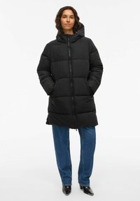 Sieviete, kas valkā melnu kapuci ar pufu, ar rok ām kabatās, zilu džinsu un melnām ar punktiem apaviem, stāv uz priekšu vērsta.