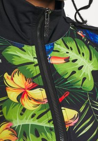 Kukkakirjoinen vetoketjullinen takki mustalla pohjalla, jossa on kirkkaan keltaisia kukkia ja vihreitä lehtiä. Yksityiskohtina korkea kaulus ja vetoketjun veto.