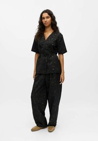 Zwarte, bloemenpatronen wikkelblouse met korte mouwen, gecombineerd met losse zwarte broeken. Het materiaal heeft een gestructureerde afwerking. Bruine slippers maken de look af.