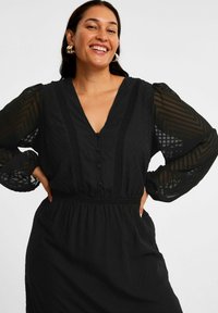 Schwarzes Kleid aus einem strukturierten, gemusterten Stoff. Mit V-Ausschnitt, Knopfverschluss und langen, durchsichtigen Ärmeln mit elastischen Bündchen. Elastischer Taillenbereich.