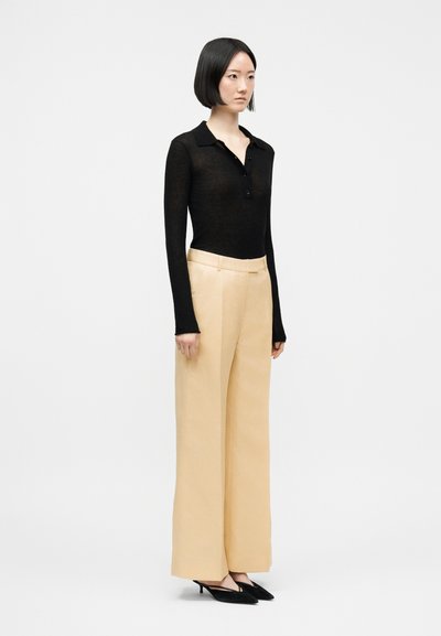 Kvinne iført svart langermet topp med krage, beige vide bukser og svarte spisse hæler, stående mot en ensfarget lys bakgrunn.