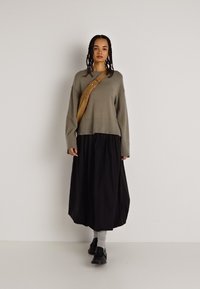 Szary sweter z dzianiny, czarna luźna spódnica, beżowa torba na ramię, szare skarpetki oraz czarne buty. Modelka stoi prosto na jednolitym tle.