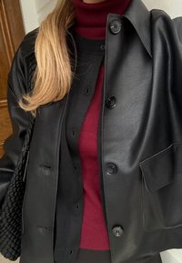 Henkilö pukeutuneena kerrospukeutumiseen: musta nahkatakki mustan neuleen ja punaisen pooloneuleen päällä, pitkät vaaleat hiukset ja musta kudottu laukku.