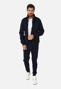 Giacca a zip blu navy e pantaloni jogger coordinati in un tessuto morbido, con un design classico e cuciture dettagliate, abbinati a sneakers bianche.