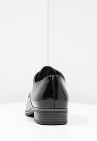 Chaussures en cuir verni noir avec un talon bas, dotées de détails de couture minimalistes et d'un bout légèrement arrondi, sur fond blanc.