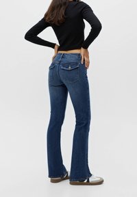 Vaqueros acampanados azul oscuro con una cintura de altura media, que presentan dos bolsillos traseros y una textura de denim suave. Combinados con una camiseta de manga larga negra.