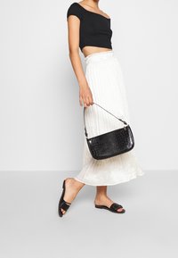 Černá kabelka s krokodýlí texturou a krátkým popruhem. Modelka má na sobě černý crop top a bílou plisé sukni. Ploché černé sandály.