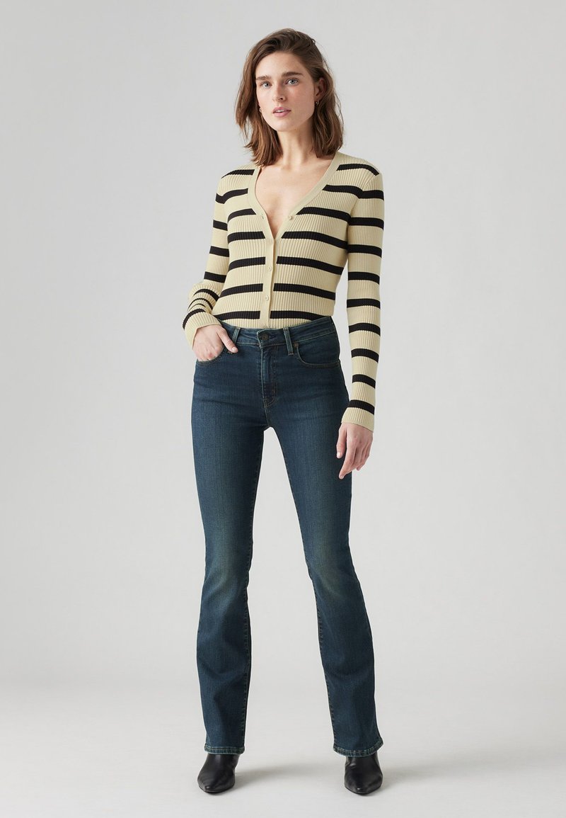 Cardigan de canalé a rayas amarillas y negras con un profundo escote en V, cierre de botones, combinado con jeans de campana oscuros y botas negras hasta el tobillo.