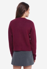 Vínový pletený svetr s kulatým výstřihem a dlouhými rukávy, zkombinovaný s šedou sukní, která má zkrácený střih a jemnou texturu.