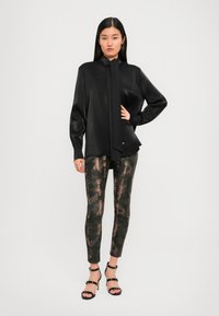 Black silk blouse with a long tie and button details, paired with form-fitting, textured leggings in green and copper tones, and black strappy heels.