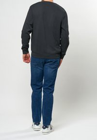Man som bär en svart långärmad sweatshirt, blå jeans och vita sneakers, stående vänd bort från kameran mot en ljus enfärgad bakgrund.