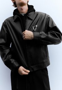 Zwarte jas van nep leer met een cropped ontwerp. Heeft een zilveren broche van kettingen en een pijl, ritssluiting aan de voorkant en een grote kraag.