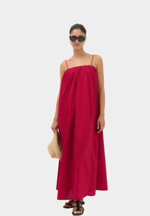 Frau trägt ein lockeres, ärmelloses rotes Maxikleid, schwarze Sandalen, dunkle Sonnenbrille, hält einen beigen gewebten Hut vor einem einfarbigen hellen Hintergrund.