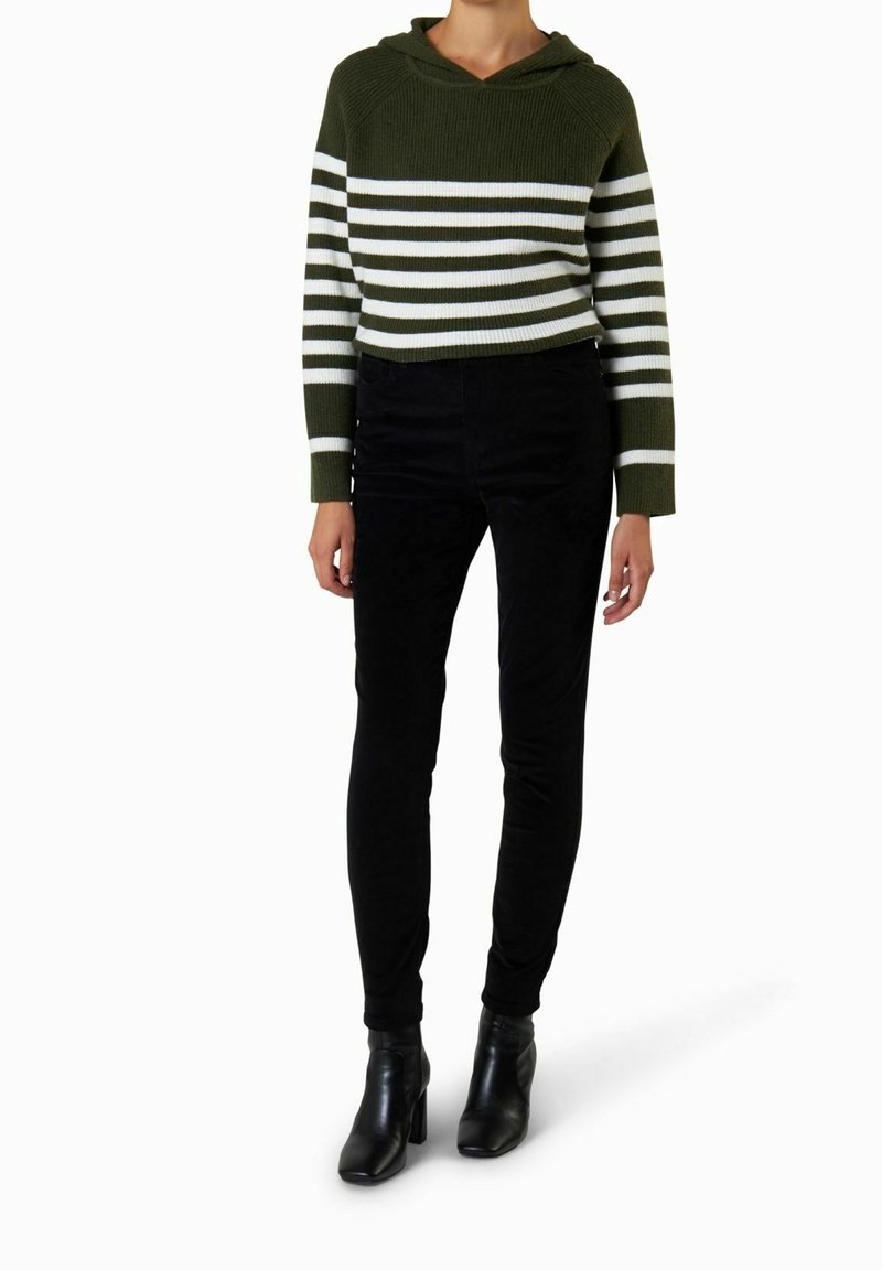 Personne portant un pull rayé vert olive et blanc, un pantalon noir ajusté et des bottines noires à talons, debout sur un fond blanc.