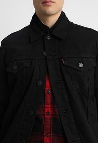 Svart denimjacka med spetsig krage, framfickor och runda knappar. Bärs över en röd rutiga skjorta med svarta knappar.