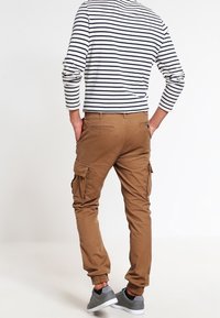 Brązowe spodnie cargo z elastycznymi mankietami, bocznymi kieszeniami i gładką fakturą. Noszone z czarną i białą bluzką z długim rękawem w paski.