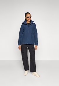 Veste imperméable bleu marine avec capuche, fermeture éclair à l'avant et logo sur la poitrine, associée à un pantalon cargo noir et des chaussures beiges.