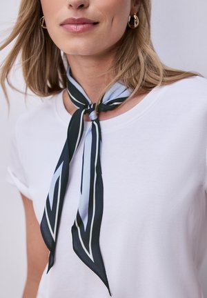 Kobieta ubrana w białą koszulkę z szalikiem w czarne i jasnoniebieskie paski zawiązanym wokół szyi oraz złotymi kolczykami kołami.