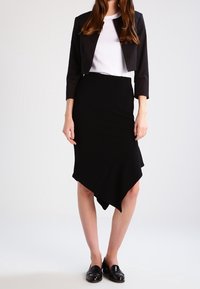 Veste noire courte portée sur une chemise blanche, associée à une jupe asymétrique noire. Coupe slim, texture lisse, couleurs unies. Moccassins noirs portés.