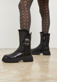 Botas de tobillo de cuero negro con un acento de hebilla, cuello de piel texturizada, cremallera lateral y suela de goma gruesa. Llevadas con medias de patrón.