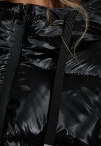 Svart puffjacka med ett glansigt, vadderat yttre. Har en frontzipper och kontrasterande svarta dragsnören för justering.