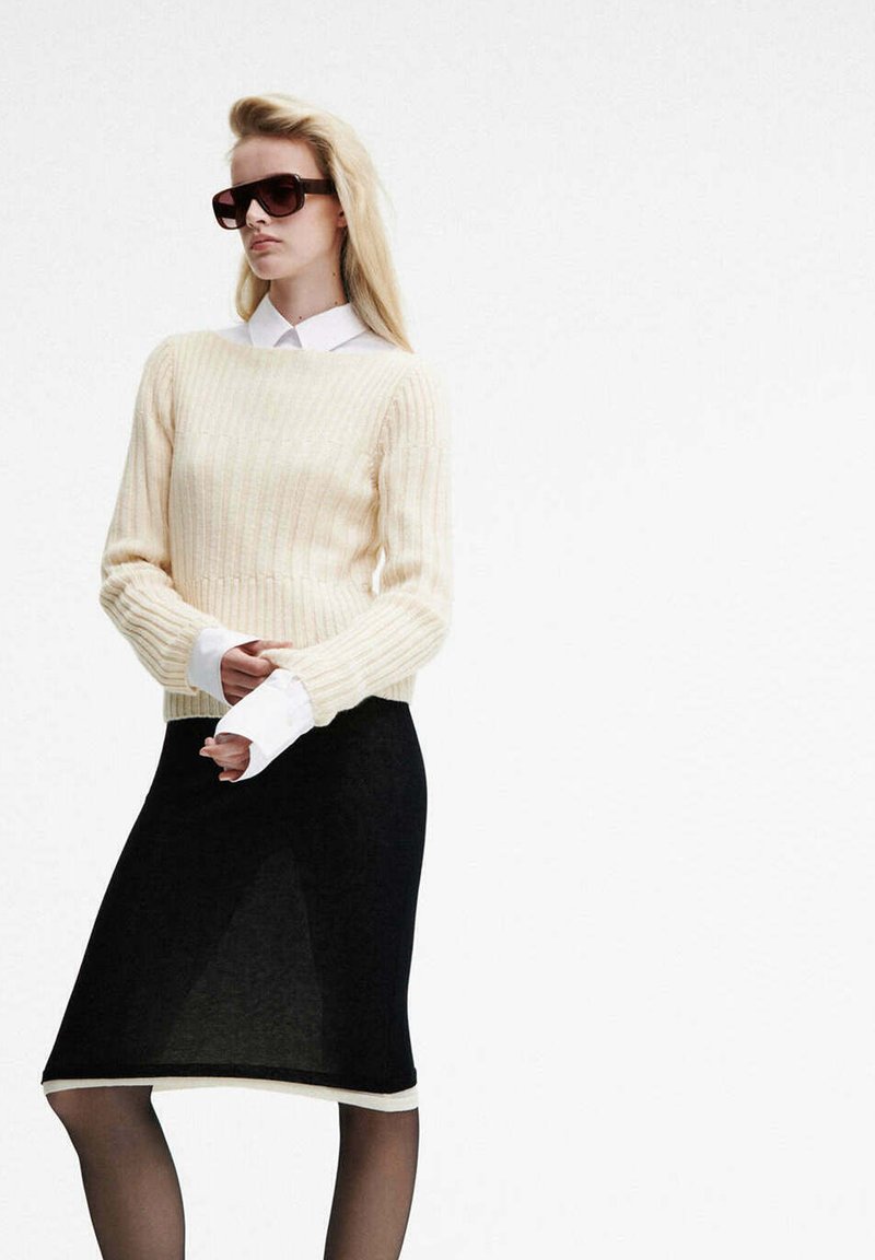 Femme portant des lunettes de soleil oversized, un pull crème sur une chemise blanche, et une jupe noire, debout devant un fond blanc uni.