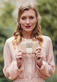 Žena s vlnitými blonďatými vlasy, oblečená v bledě růžové blůze, drží béžovou peněženku s bílým popruhem venku.