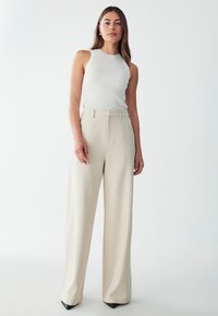 White ribbed tank top paired with high-waisted, wide-leg beige trousers. Model wears black pointed heels. Simple, clean design and fit.
