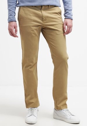 Homme portant un pantalon beige, une chemise à manches longues bleu clair et des baskets blanches, debout devant un fond uni.
