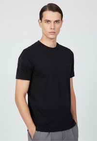 Svart t-shirt med kort ärm i slät tyg, med texturiserade detaljer på axlarna och en subtil logotyp på bröstet.