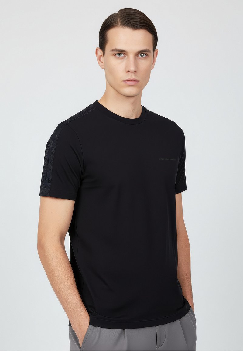 Svart t-shirt med kort ärm i slät tyg, med texturiserade detaljer på axlarna och en subtil logotyp på bröstet.