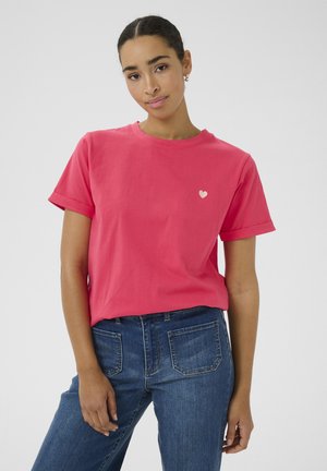T-shirt di cotone rosa con maniche corte arrotolate, caratterizzata da un piccolo logo a forma di cuore bianco sul petto. Abbinata a jeans in denim blu.
