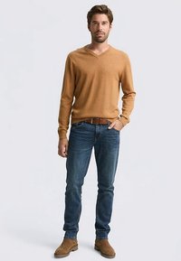 Sweter z dekoltem w serek w kolorze camel, wykonany z miękkiego materiału; noszony na niebieskie dżinsy. Brązowy pasek i jasnobrązowe botki za kostkę. Modelka stoi na jasnym tle.