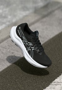 Zwarte hardloopschoen met een gestructureerde bovenkant, witte accenten en een dikke witte zool. Heeft Asics-logo en een ademend ontwerp.
