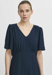 Navy blue dress with short puff sleeves, V-neckline, textured fabric, and gathered waistline details. Subtle dot pattern throughout.