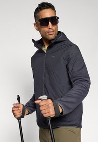 Veste zippée noire avec capuche, fabriquée en matériau lisse. Le modèle tient des bâtons de randonnée, portant de grandes lunettes de soleil noires sur un fond neutre.