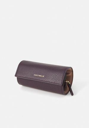 Portafoglio in pelle di colore viola con texture, chiusura a patta, logo dorato "Coccinelle" e tasca con zip sul lato. Forma cilindrica compatta.