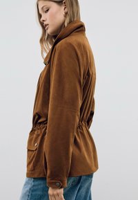 Jeune femme portant une veste en daim brun avec une taille cintrée et une poche à rabat, vêtue d'un jean bleu, se tenant contre un fond clair.
