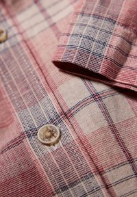 Rode en blauwe geblokte katoenen shirt met een knoop. Heeft een gestructureerde weving, opgevouwen manchet en een lichte beige achtergrond.