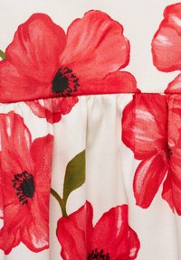 White fabric with large red poppy flowers, featuring black centers and green leaves, with a gathered texture along the seam.