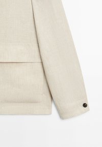 Beige linen jacket sleeve with a black button on the cuff and a large patch pocket on the side.