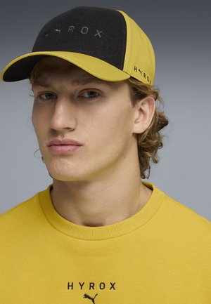 Giovane uomo che indossa un berretto giallo e nero e una felpa gialla con il logo "HYROX" e Puma sul davanti, sfondo neutro.