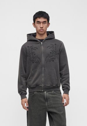 Jeune homme portant un sweat à capuche zippé gris foncé avec des motifs brodés abstraits noirs et un jean gris foncé, debout devant un fond uni.