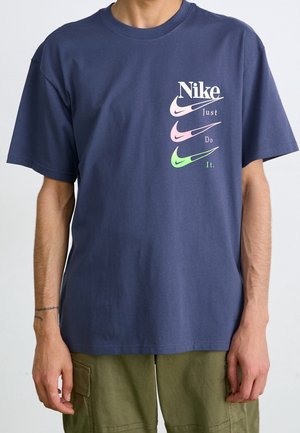Homme portant un t-shirt Nike bleu marine avec trois logos swoosh multicolores et le texte "Just Do It." sur la poitrine gauche, associé à un pantalon vert olive.