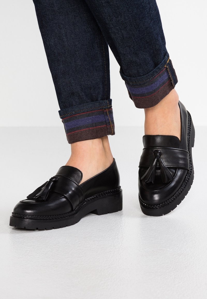 Mocassins à pampilles en cuir noir portés avec un jean foncé retroussé sur un sol blanc lisse.