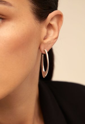 Pendiente de aro plateado con una forma elongateda y abstracta. Textura suave, acabado pulido y una abertura estrecha en un lado.