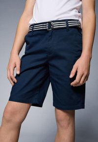 Tengerészkék rövidnadrág sima textúrával, elülső zsebekkel, fekete-fehér csíkos övvel, amelyen ezüst csat található.