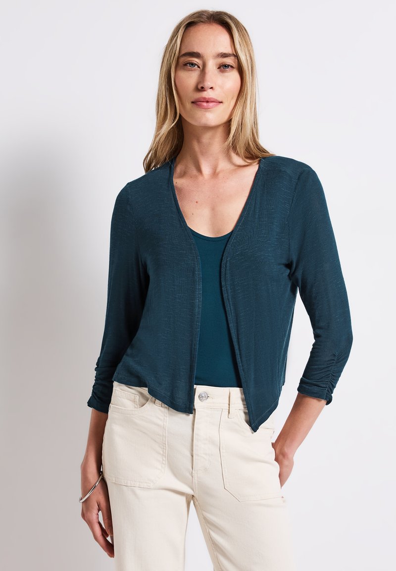 Teal, lätt cardigan med trekvartsärmar och öppen framsidan, parat med en mörk topp och beige byxor. Slät textur.