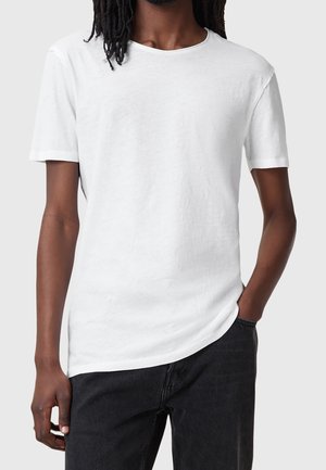 Mand med mørk hud iført en ensfarvet hvid kortærmet t-shirt og sorte bukser, hånd i lommen, foran en lysegrå baggrund.