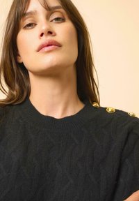 Czarny sweter w splot kablowy z okrągłym dekoltem i krótkimi rękawami. Zawiera charakterystyczne złote guziki na ramieniu.